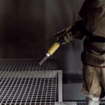 Facharbeiter mit Schutzanzug verwendet Sandstrahlschlauch in geschlossener Strahlkabine zur präzisen Reinigung von Metallteilen