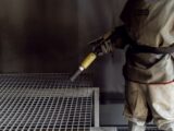 Facharbeiter mit Schutzanzug verwendet Sandstrahlschlauch in geschlossener Strahlkabine zur präzisen Reinigung von Metallteilen